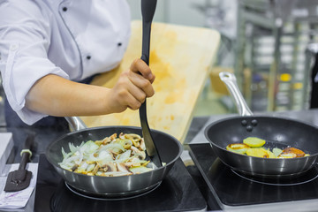 Chef hand cooking sliced champignon mushrooms and potatoes in frying pans with oil on electric stove at cuisine of restaurant. Professional cooking, catering, cookery, gastronomy and food concept