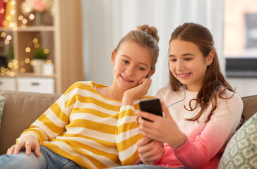 people, technology and friendship concept - happy teenage girls with smartphone and earphones listening to music sitting on sofa at home