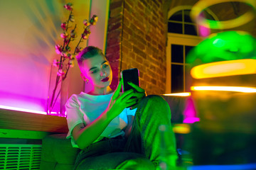 Obraz premium Selfie. Cinematic portrait of stylish woman in neon lighted interior. Toned like cinema effects, bright neoned colors. Caucasian model using smartphone in colorful lights indoors. Youth culture.