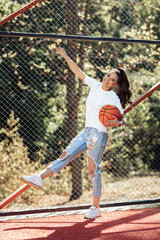 Adorable brunette young woman with bright smile dressed in casual modern clothes posing with basketball at the stadium.