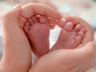 Baby feet in mother hands. Tiny Newborn Baby's feet on female Heart Shaped hands closeup. Mom and her Child. Happy Family concept. Beautiful conceptual photo of Maternity.
