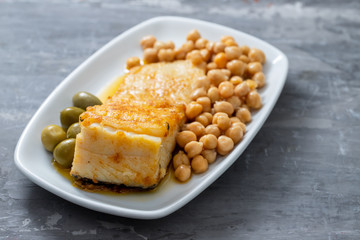 fried cod fish with chick pea and olives on white dish on ceramic background