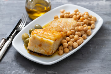 fried cod fish with chick pea and olives on white dish on ceramic background