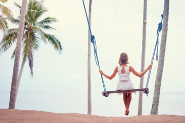 Vacation concept. Happy young woman in white dress sitting on swing enjoying sea view.