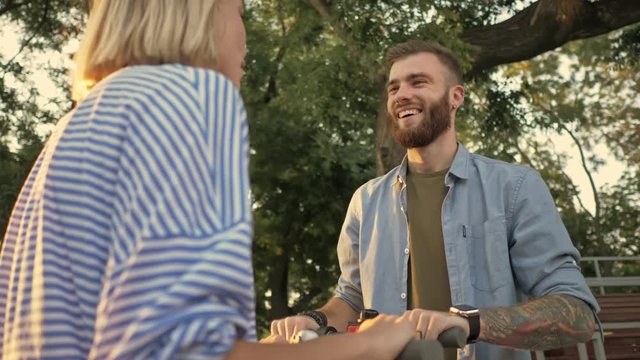 Happy young friends guy and girl are laughing and talking while driving electric kick scooters at green park landscape at sunset time