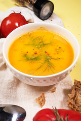 Close-up white bowl with vegetable cream soup on beige kitchen napkin with tomatoes, bread, spoon and spices. Vertical.