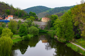 Fototapeta na wymiar Reflections in the water of Ohre river between trees in Loket, Czech Republic, with typical houses, a stone wall and a bridge at the background