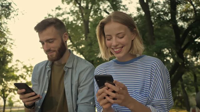 Young smiling couple is using smartphones while sitting on the bench at a park landscape