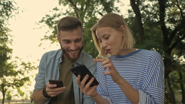 Young smiling girl is showing something on her smartphone to her boyfriend while sitting on the bench at a park landscape