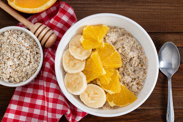 Bowl of oatmeal porridge with banana and orange  on wooden background. Top view, copy space