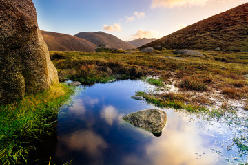Puddle of water with reflection of blue sky and clouds on the Hares Gap. Big boulders at Mourne Mountains range and beautiful valley, Northern Ireland