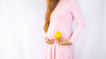 A close up photo of a pregnant young woman holding a lemon next to her belly showing the size of the baby in a beautiful dress. Photos of fetal growth at 14 weeks pregnancy. Healthy pregnancy diet.