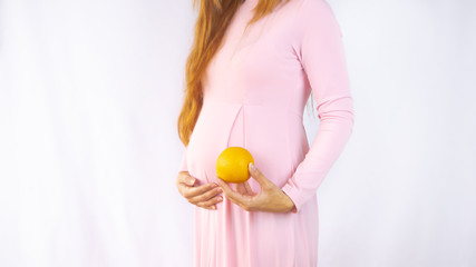 A close up photo of a pregnant young woman holding an orange next to her belly showing the size of the baby in a beautiful dress. Photos of fetal growth 15 week pregnancy. Healthy pregnancy diet