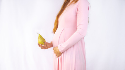 A close up photo of a pregnant young woman holding a pear next to her belly showing the size of the...
