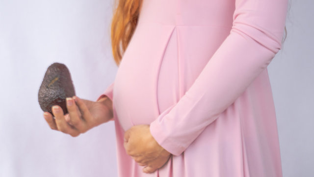 A Close Up Photo Of A Pregnant Young Woman Holding A Avocado Next To Her Belly Showing The Size Of The Baby In A Beautiful Dress. Photos Of Fetal Growth At 16 Weeks Pregnancy. Healthy Diet For Weight.