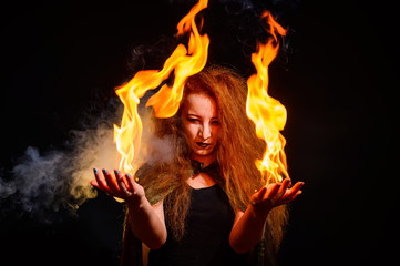 An ominous witch with long curly hair holds a magical fire. Tongues of flame on the palms of a...