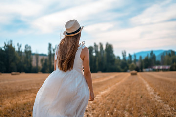 Lifestyle, young blonde long-haired peasant with long white dress and a hat in a dry straw field