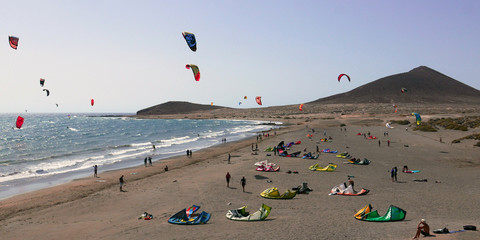 Many colorful kites on beach and kite surfers riding waves and flying during windy day in canarian El Médano in Tenerife with Montana roja hills on horizon © Petr