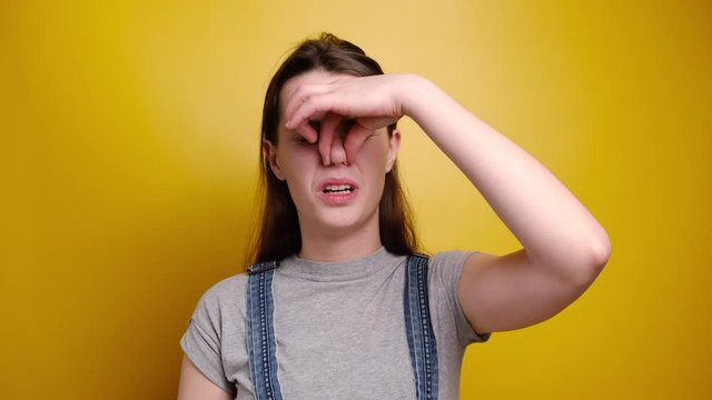 Bad odour concept. Discontent brunette girl covers nose with hand, smells something awful, pinches nose, frowns in displeasure, dressed in grey t-shirt, isolated over yellow background.