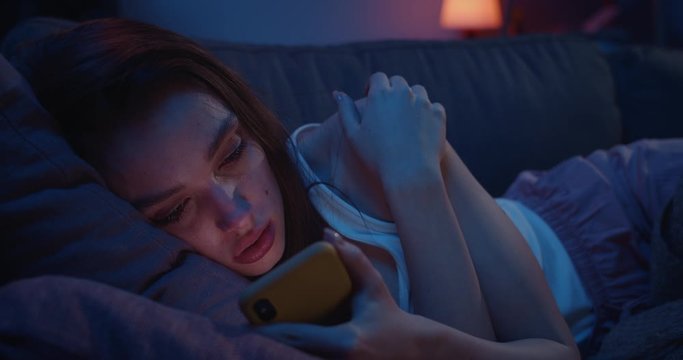 Close up of unhappy young woman lying on sofa and staring at smartphone screen. Millennial female feeling depressed and lonely after painful brakeup and using mobile phone.