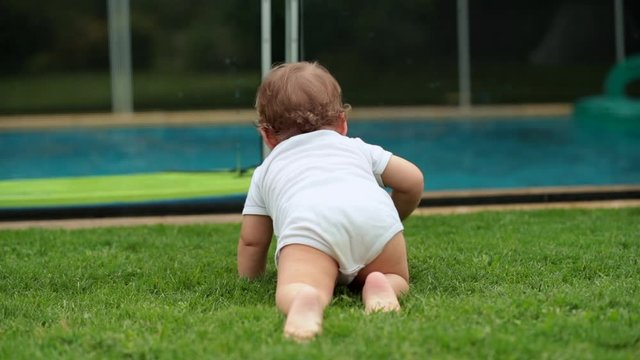 Baby crawling in backyard grass observing children play inside pool. Infant crawls outside