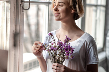 Smiling young woman near the window holds a bouquet of purple flowers near her face 