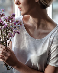 Smiling young woman near the window holds a bouquet of purple flowers near her face  - 324882219