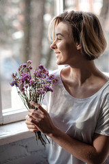Smiling young woman near the window holds a bouquet of purple flowers near her face  - 324882242