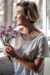 Smiling young woman near the window holds a bouquet of purple flowers near her face  - 324882243
