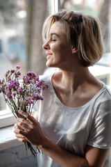 Smiling young woman near the window holds a bouquet of purple flowers near her face  - 324882265