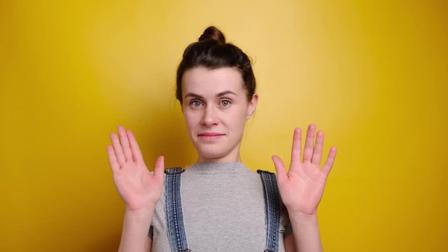 Attractive brunette female shows refusal gesture, doesn`t want to participate in meeting, leave me in piece, wears grey t-shirt and denim overalls, poses against yellow  studio background