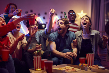 Group of young fans sitting on sofa drinking beer and eating pizza while watching football match on TV at home