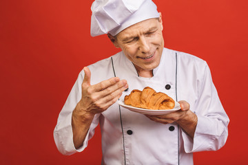 A portrait of a senior baker holding croissant isolated on red background.