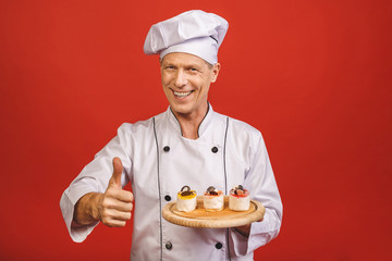 A portrait of a senior baker holding delicious cakes isolated on red background. Thumbs up.