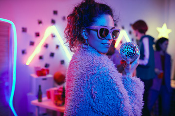 Portrait of young party girl in sunglasses and in stylish jacket looking at camera while dancing at...