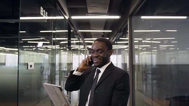 Attractive smiling professional black-skinned office manager standing in long company corridor and talking on phone