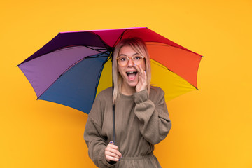 Young blonde woman holding an umbrella over isolated yellow wall shouting with mouth wide open