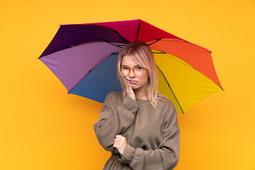 Young blonde woman holding an umbrella over isolated yellow wall unhappy and frustrated