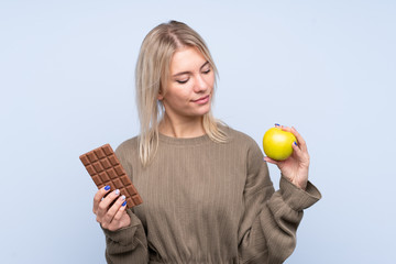 Young blonde woman over isolated blue background taking a chocolate tablet in one hand and an apple...