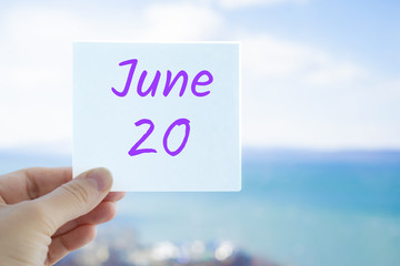 June 20th. Hand holding sticker with text June 20 on the blurred background of the sea and sky. Copy space for text. Month in calendar concept