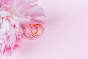 A pair of gold wedding rings on pink peony flower petals on a light pink background with a copy space