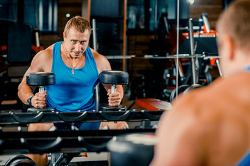 Young muscular guy doing hard exercises with dumbbells on the training bench in the gym