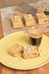 Top, front view, close distance of a torn open, Apricot Pecan Bars on a yellow, round plate with a cup of espresso and three bars cooling on parchment paper on a wood cutting board
