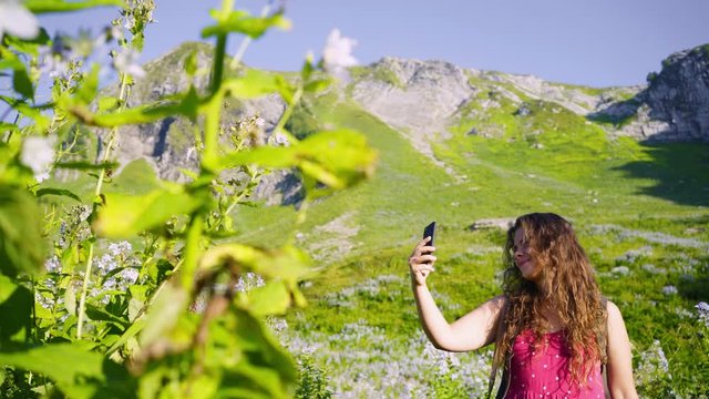 Pretty girl traveller taking selfie in mountain. Carefree woman in pink sundress posing, looking at camera while taking selfie on smartphone among blooming plants in sunny day