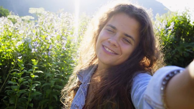 Pretty girl traveller taking selfie in mountain. Close-up portrait woman in bright backlit posing, looking at camera while taking POV selfie among blooming field in sunny day
