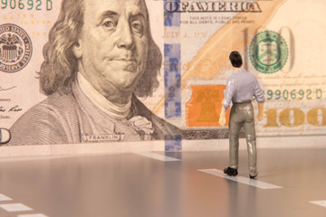 A male miniature figure walking toward a US 100 dollar bill