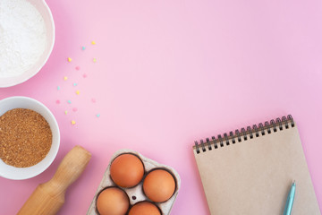 Easter baking ingredients and notebook on light pink background. Eggs, flour, rolling pin, sugar and colorful confetti on table. Cooking concept and holiday. Top view, flat lay, copy space