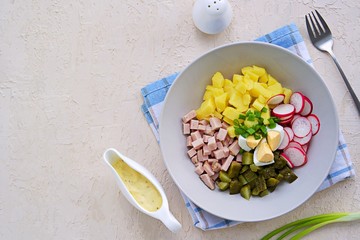 Potato salad with ham, fresh radish and pickled cucumber in a gray bowl on a light concrete background. American cuisine. Potato Recipes. Top view, copyspace