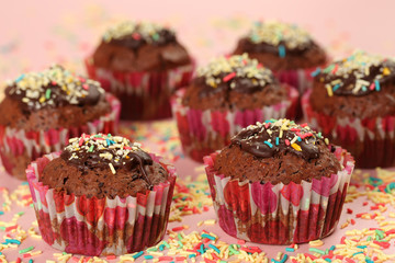 Homemade, delicious cocoa muffins with dark chocolate icing and multicolored sugar sprinkles on a pink background. Selective focus, shallow depth of field