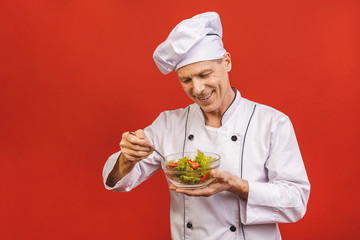 Picture of happy young senior chief cook in uniform standing isolated over red wall background, holding salad.